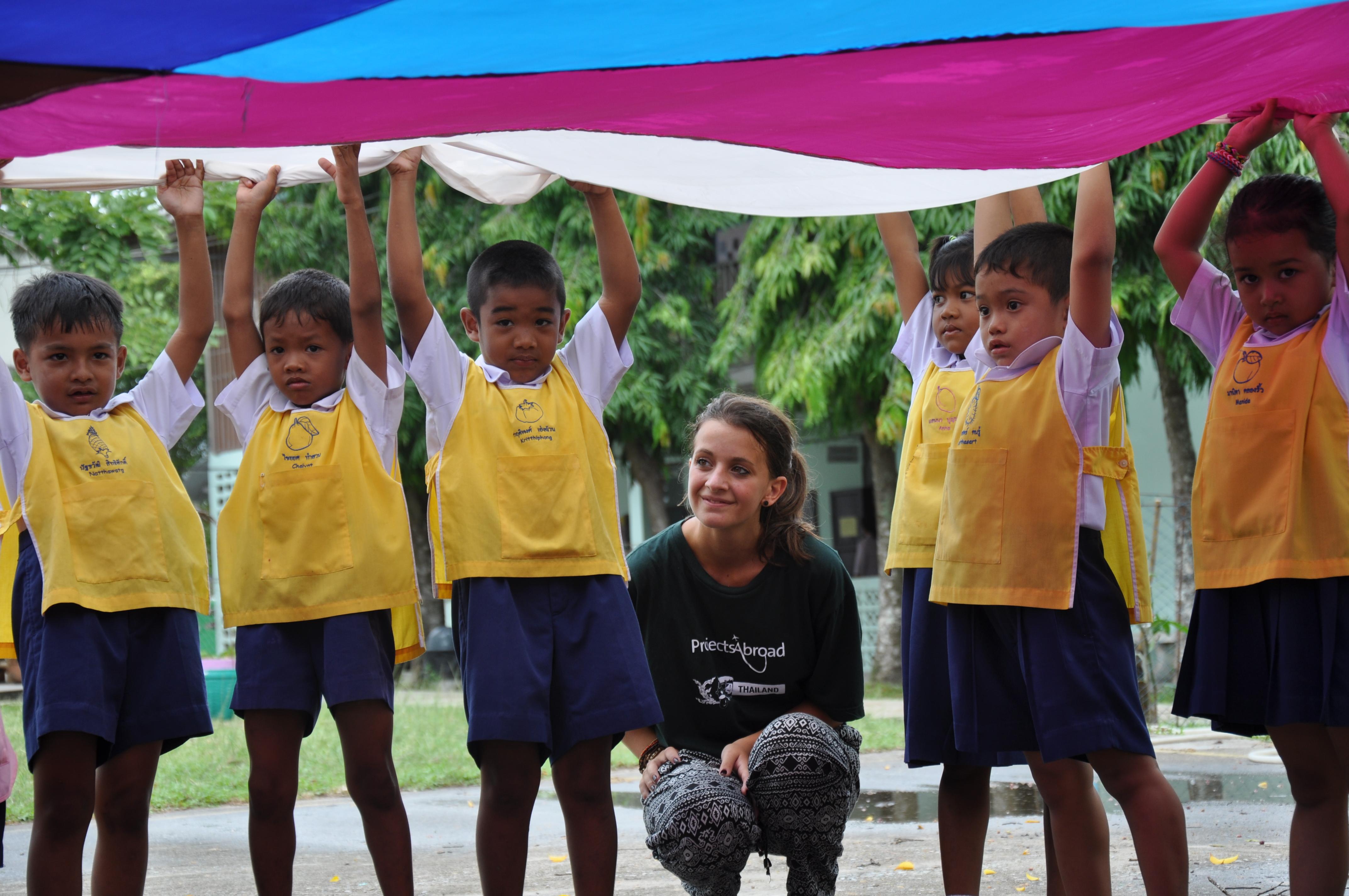 Projects Abroad volunteer under a parachute with children during an outdoor activity at a kindergarten in Thailand. A volunteer working with children in Thailand crouches under a parachute during an activity with kids at her Childcare placement.